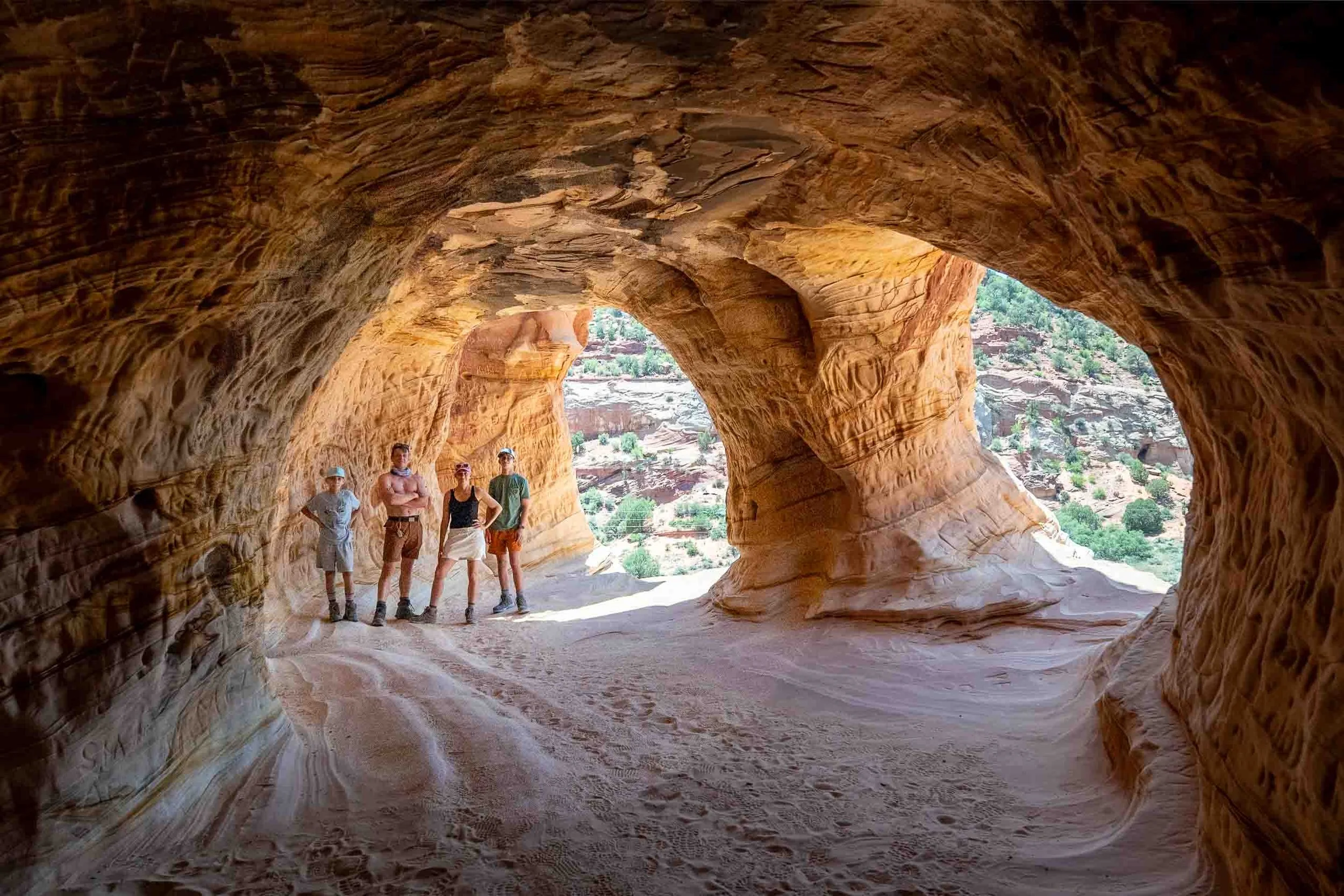 Four people standing in a sandstone cave with natural arches and a canyon landscape visible in the background.