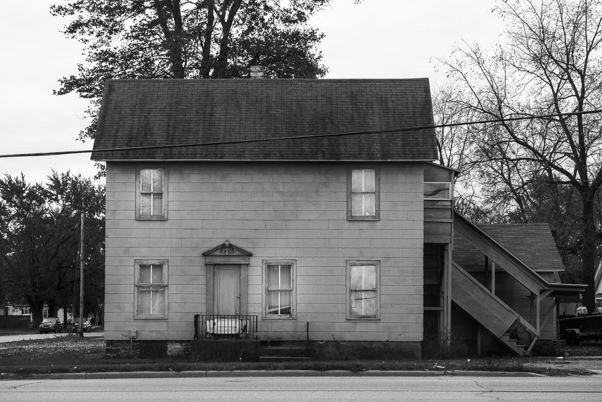 A two-story house with a steep roof, boarded-up front door, and boarded windows. The house is in black and white and appears to be abandoned, with a staircase on the right side leading to an upper entrance.