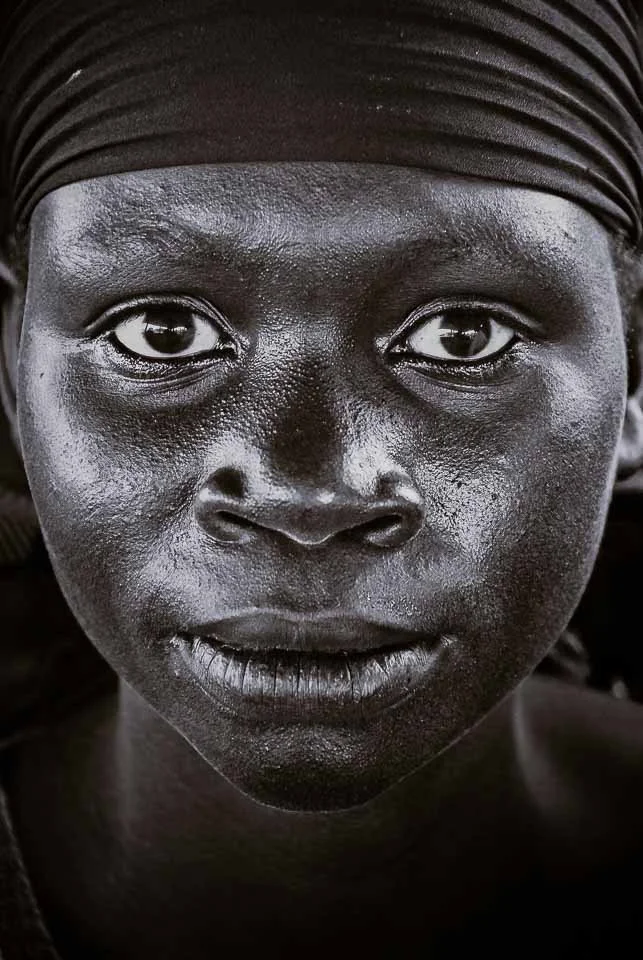 Close-up of a woman with dark skin wearing a headwrap, looking directly at the camera with a serious expression.