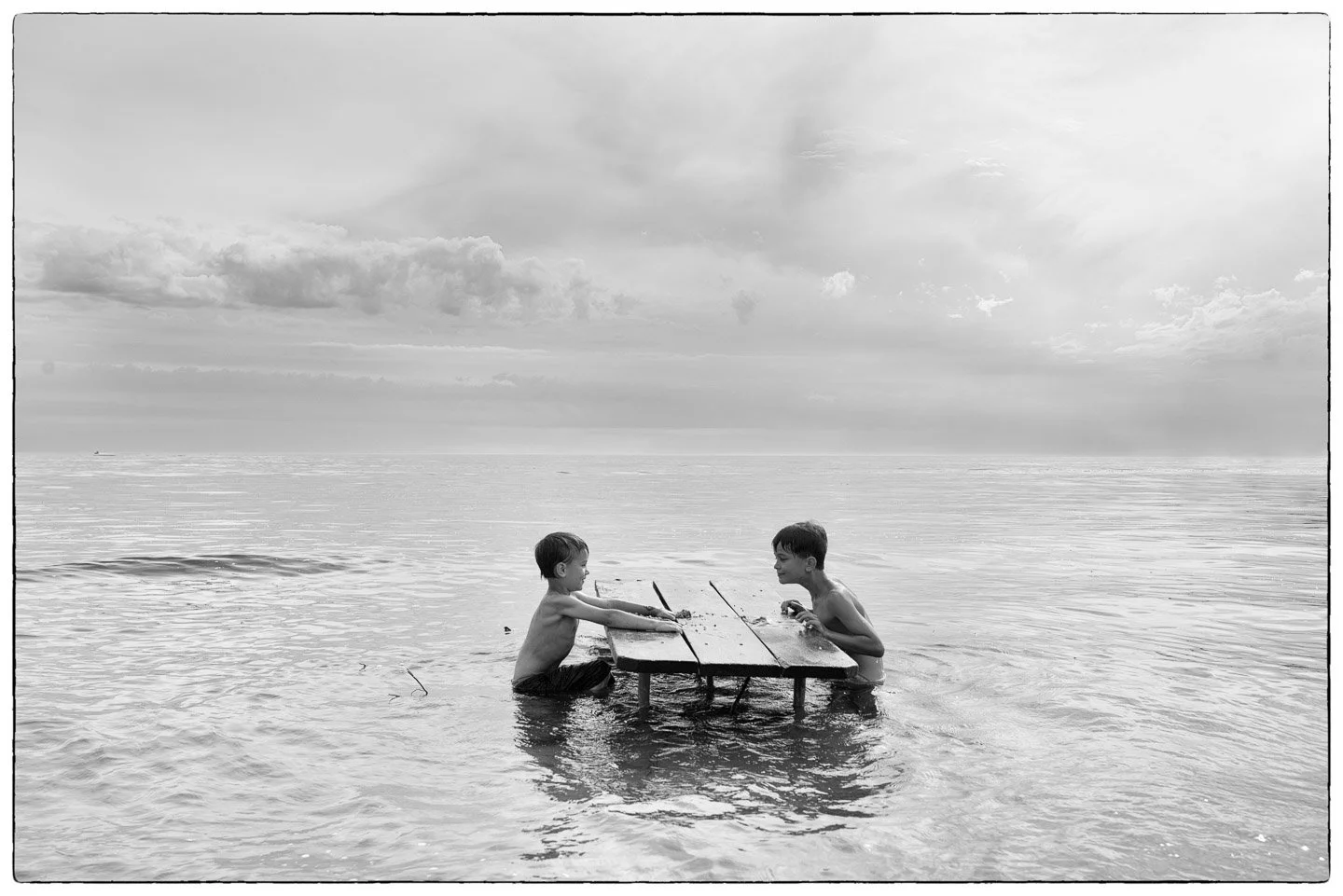 Two young boys playing a game of chess on a small wooden table while half submerged in water at the beach, with an ocean and cloudy sky in the background.