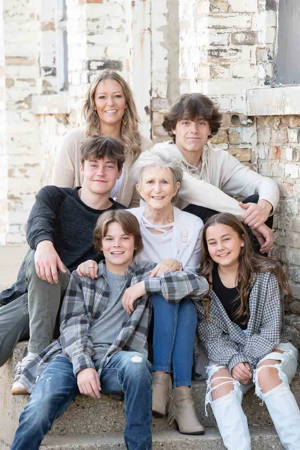 A smiling multigenerational family of seven people, including an elderly woman, two adult women, and four children, sitting and standing against a brick wall.
