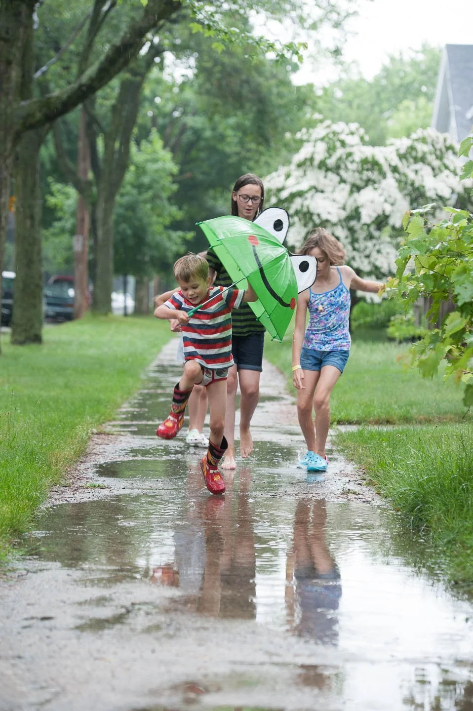 Three children, two girls and one boy, walking on a wet sidewalk during the rain with a green frog-shaped umbrella, surrounded by green trees and bushes.