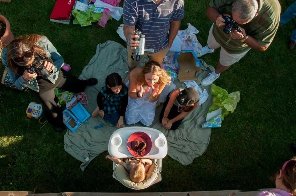 A birthday celebration outdoors with a group of people, including children and adults, gathered around a table with a cake, opening gifts, and taking photos on a grassy area.