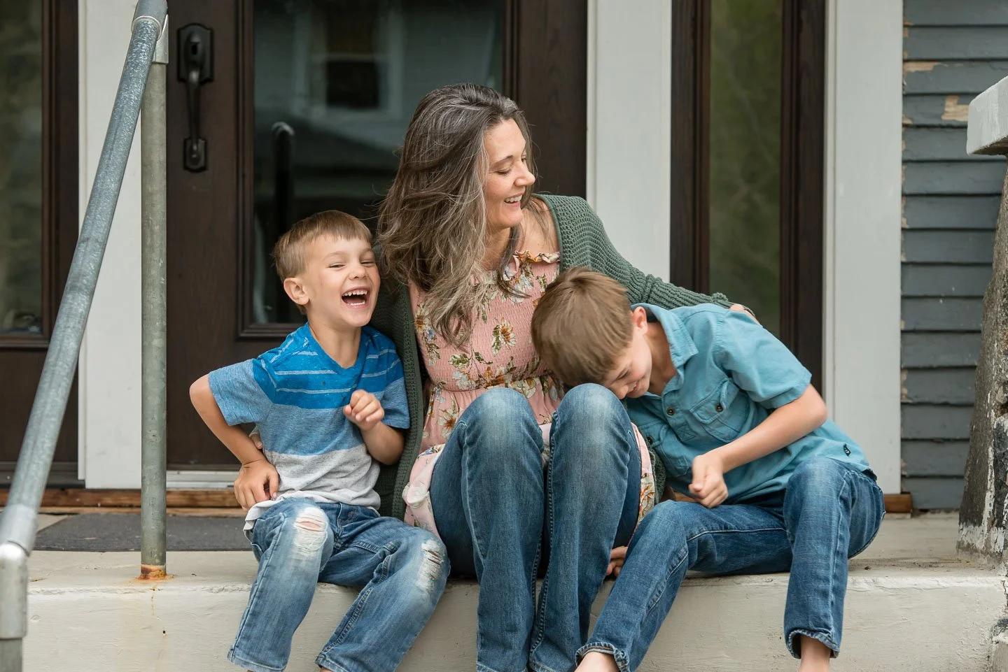 A woman and two young boys sitting on the front steps of a house, laughing and enjoying each other's company.