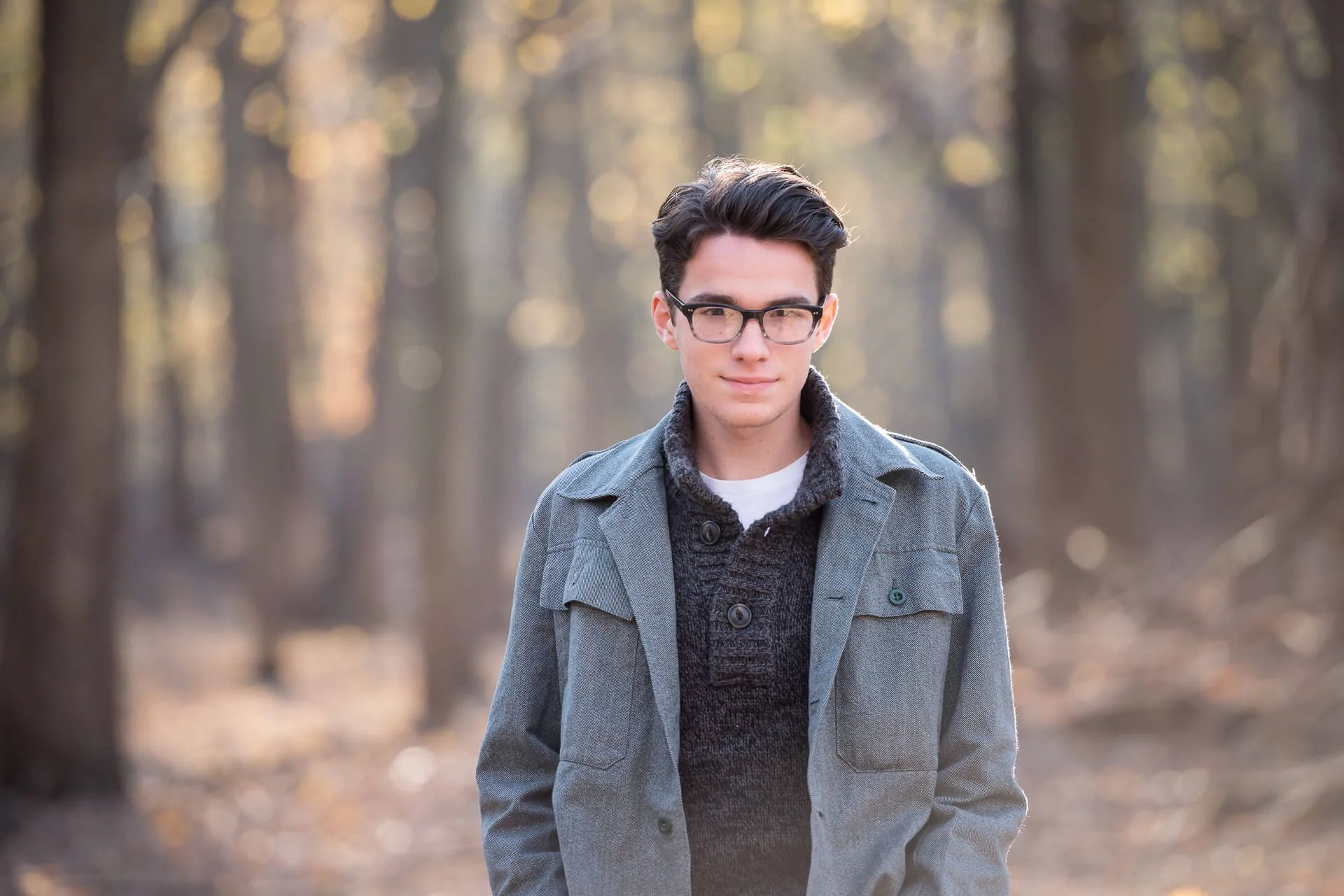 A young man with dark hair, glasses, and fair skin standing in a forest, wearing a gray jacket over a dark sweater.