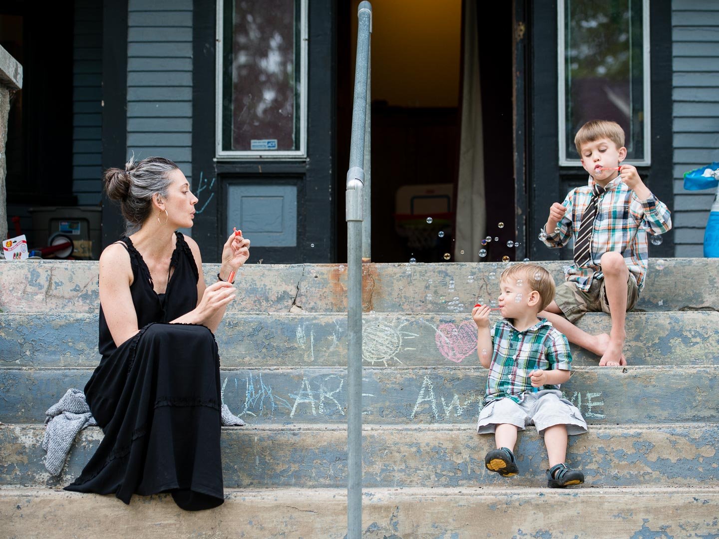 A woman and two young boys sitting on concrete steps outside a house, playing with bubbles on a sunny day.