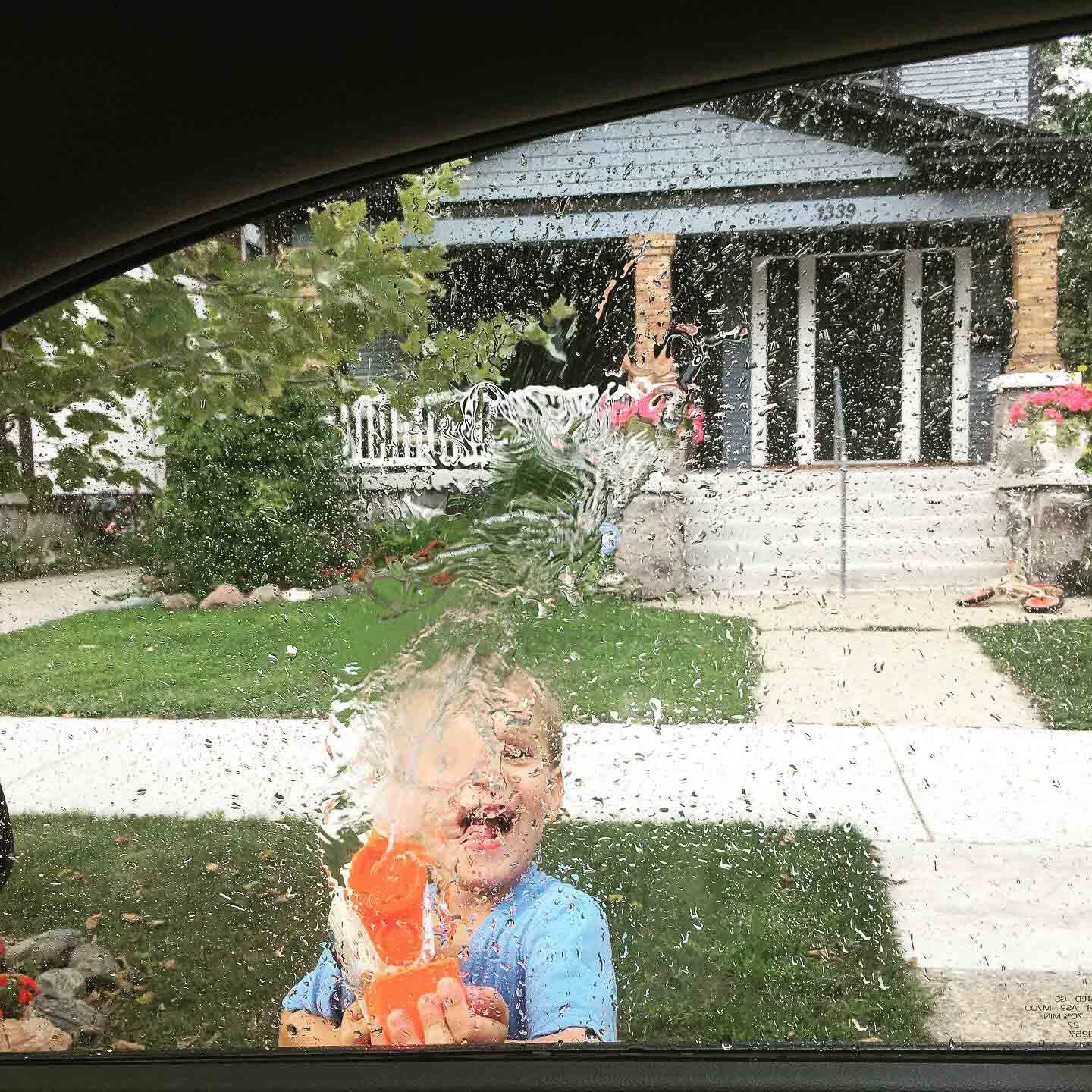 A young child is outside behind a rain-covered window, holding a water gun and smiling.