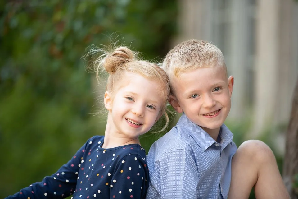 A young girl and boy smiling outdoors, leaning their heads together.