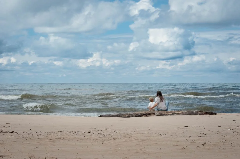 A woman and a child sitting on a log on the beach, facing the ocean with cloudy sky.