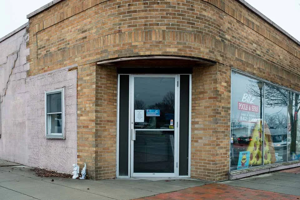 Brick building with a glass door and a window displaying a pizza shop sign, located on a sidewalk.