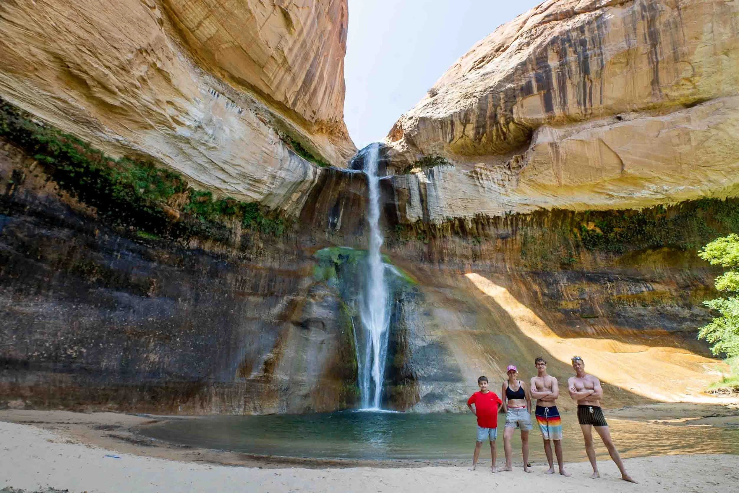 Four people standing on a beach in front of a waterfall and rock canyon, with blue sky overhead.