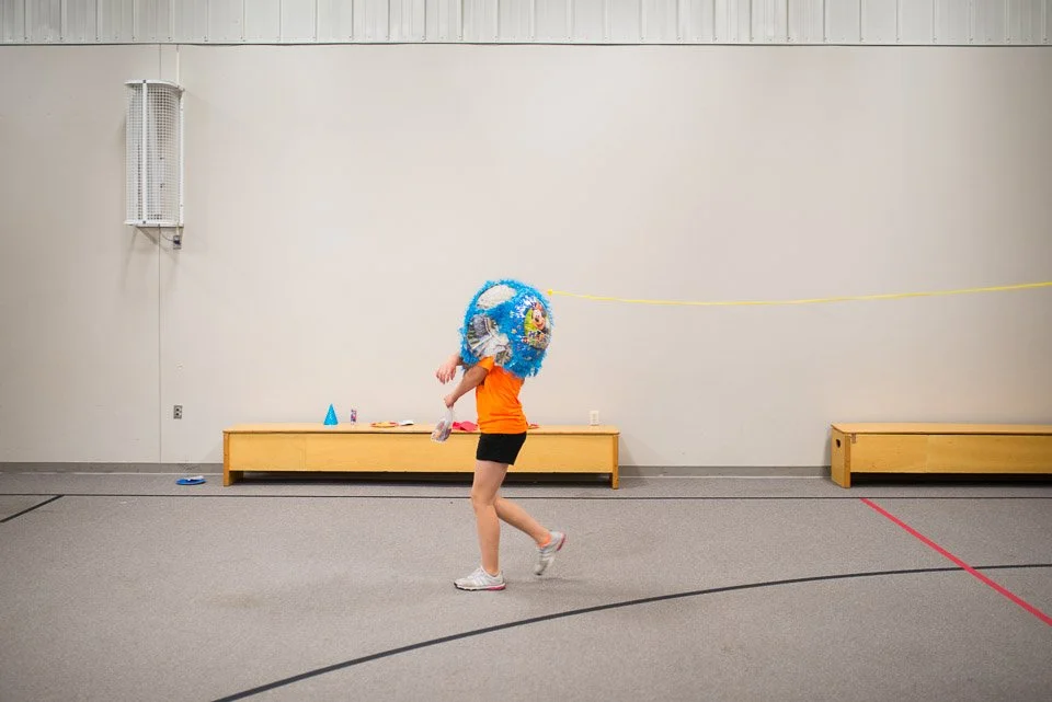A person in an orange shirt and black shorts walking in an indoor gym or recreation room, carrying a large, decorated piñata shaped like a blue ball with a cartoon character on it.