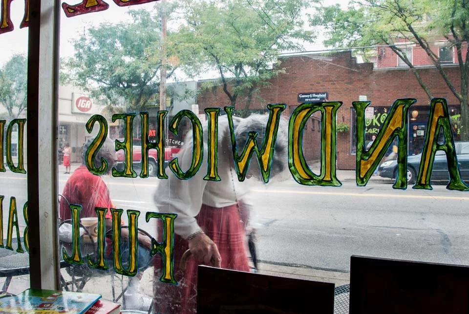 View of a street outside through a storefront window with painted green and yellow letters reading "Andwiche" and part of "Full", with two people, one standing outside and the other seated inside, visible through the window.