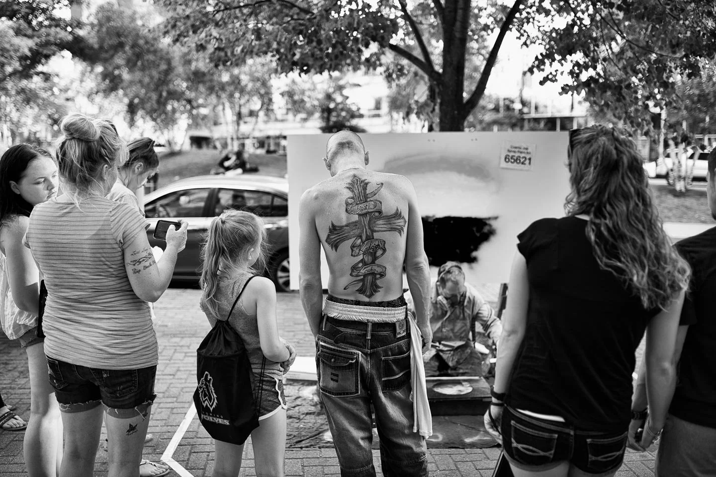 A black and white photo of a group of people gathered around a street artist who is painting a graffiti-style artwork on a canvas outdoors. The artist is shirtless, showing a large tattoo on his back, and is sitting on a stool. The onlookers include 