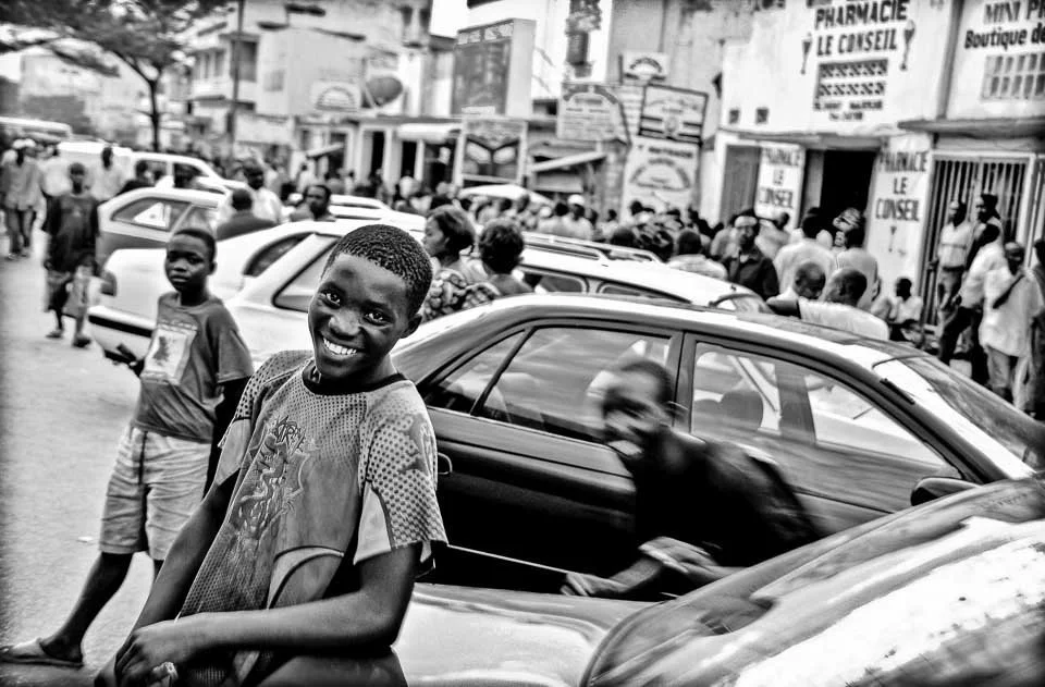 A young boy smiling at the camera on a busy street with cars and people in the background.