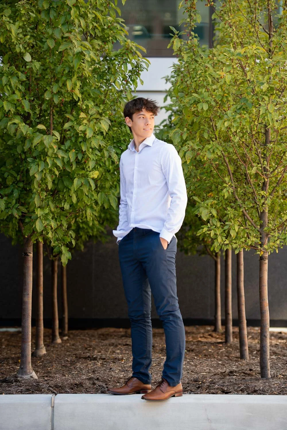 A young man with dark hair in a white dress shirt and dark pants stands outdoors with hands in pockets, surrounded by green trees.