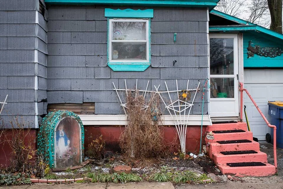 Front of a house with gray shingle siding, a small window with teal trim, and red stairs leading to a white door. Decorations include a painted curbside mirror surrounded by teal dots, a dried plant, and a metal spider web ornament. There is a blue t