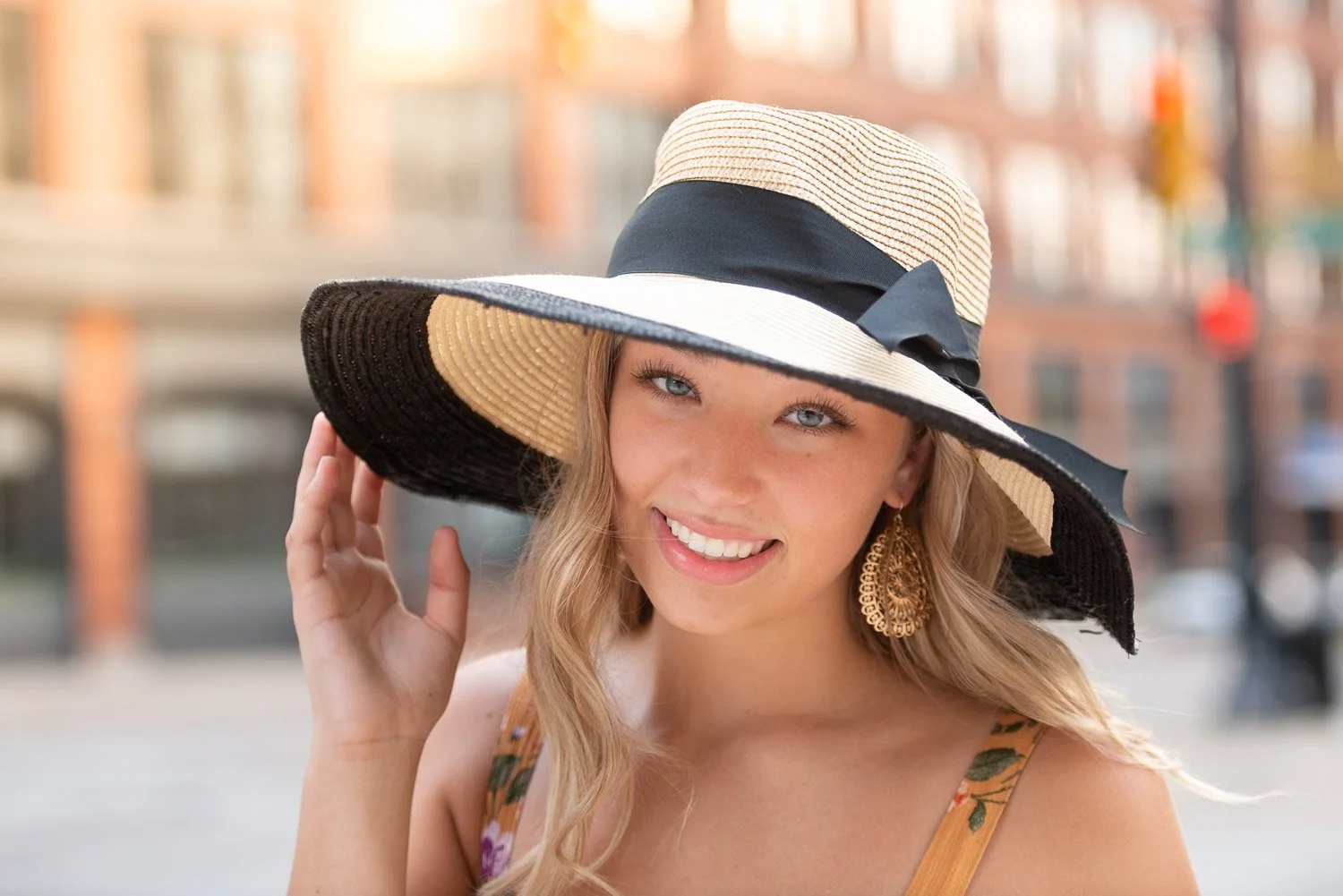 A young woman with blonde hair wearing a large straw sun hat with a black ribbon, smiling outdoors.