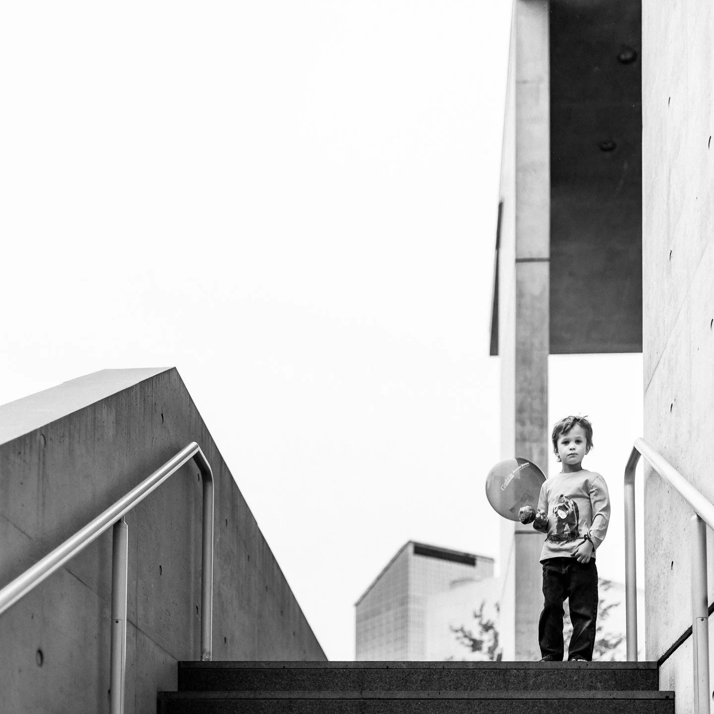 A young child holding a balloon and a toy walking up outdoor stairs between concrete walls, with modern buildings in the background.