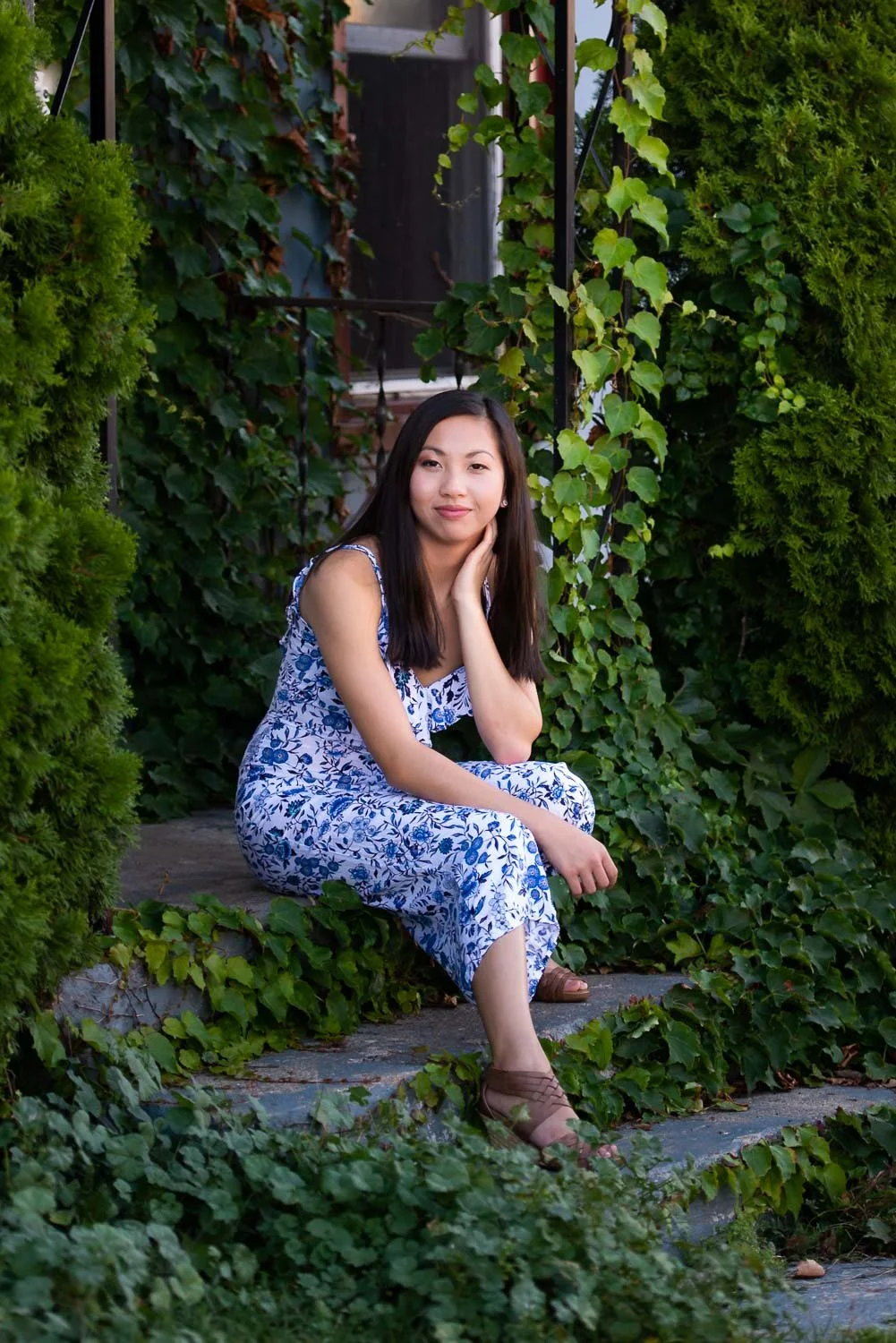 A young woman with long dark hair wearing a white and blue floral dress, sitting on a stone step surrounded by green ivy and bushes, with her hand resting on her face, outdoors in a garden.