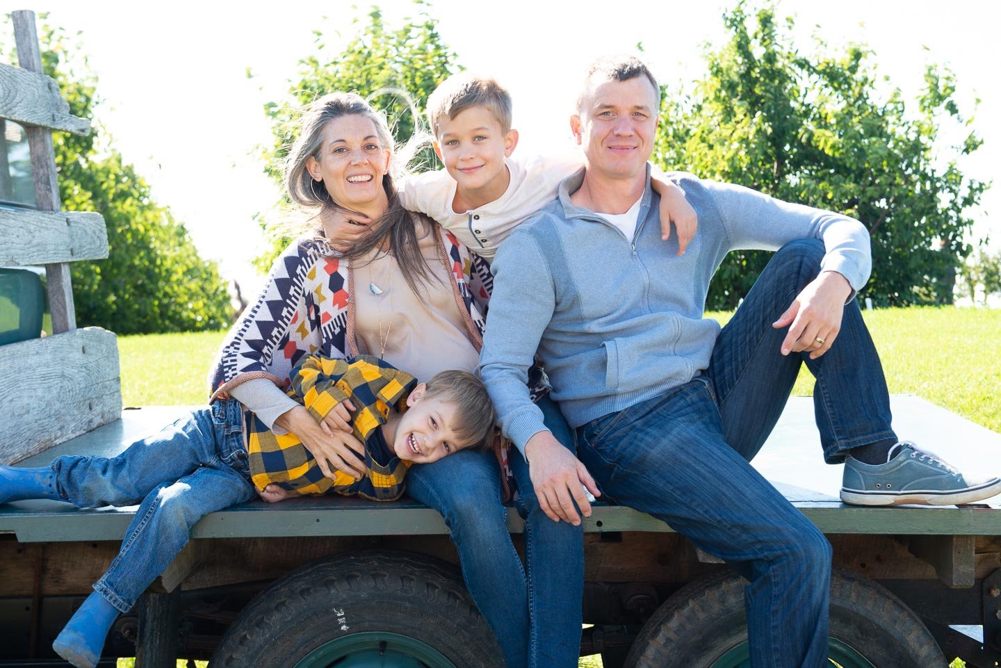 A family of four sitting on a wooden flatbed truck outdoors. The mother, father, and their two children are smiling. The mother has long gray hair and is wearing a patterned cardigan; the father has short hair and is wearing a light blue zip-up jacke