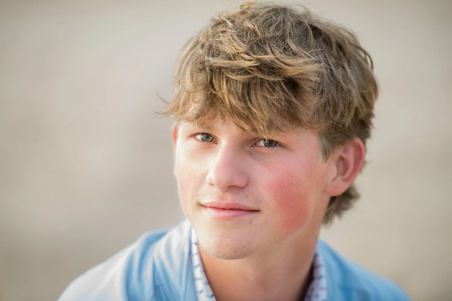 A young man with light brown, tousled hair and blue eyes, looking at the camera with a slight smile, outdoors with a blurred neutral background.