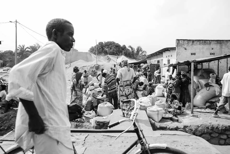 A busy outdoor market scene with people shopping and selling goods, some sitting and some standing, with makeshift stalls and sacks of items visible.