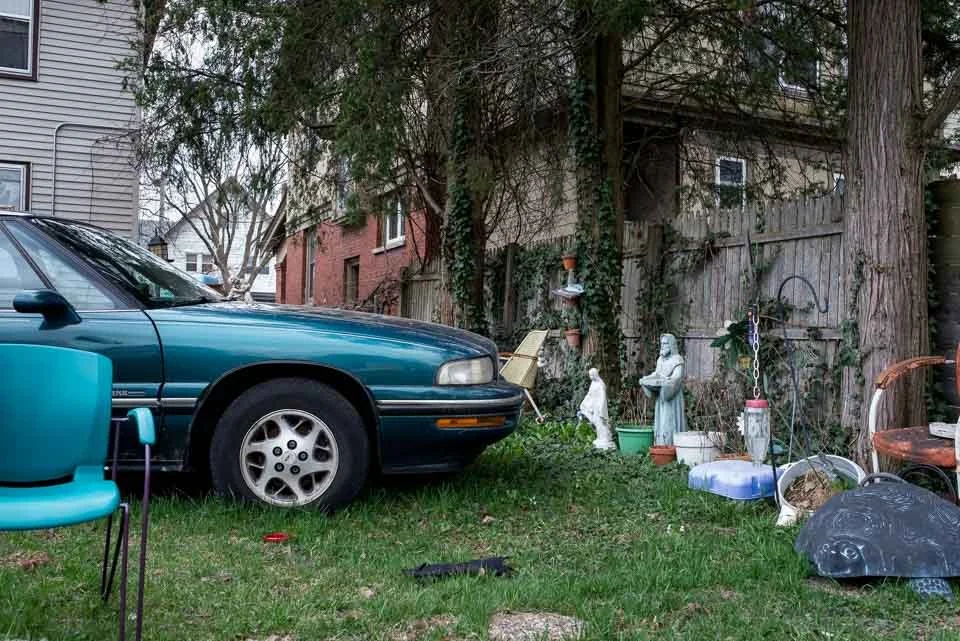 A teal-colored car parked in a grassy backyard next to a wooden fence, with garden statues, potted plants, and outdoor furniture around.
