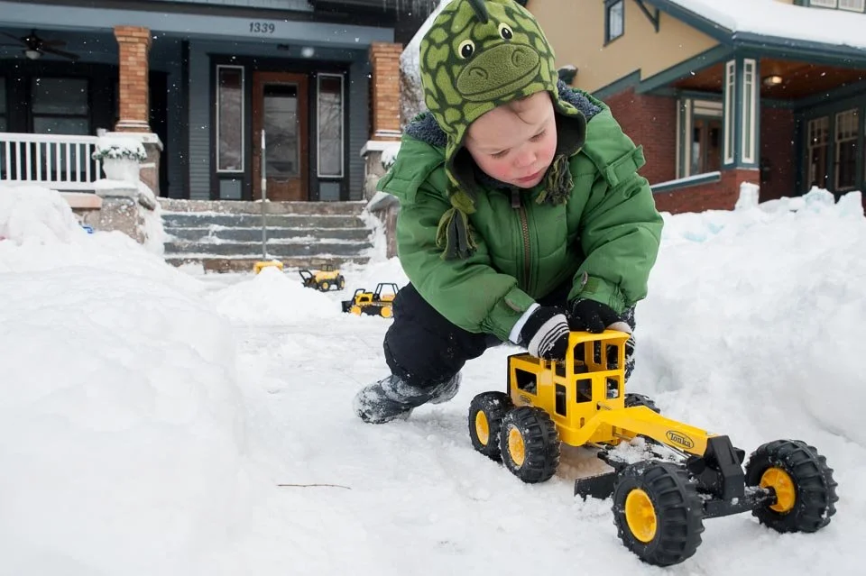 A young boy wearing a green jacket and a dinosaur hat playing in the snow, pushing a yellow toy bulldozer in front of a house with stairs and other toy vehicles nearby.
