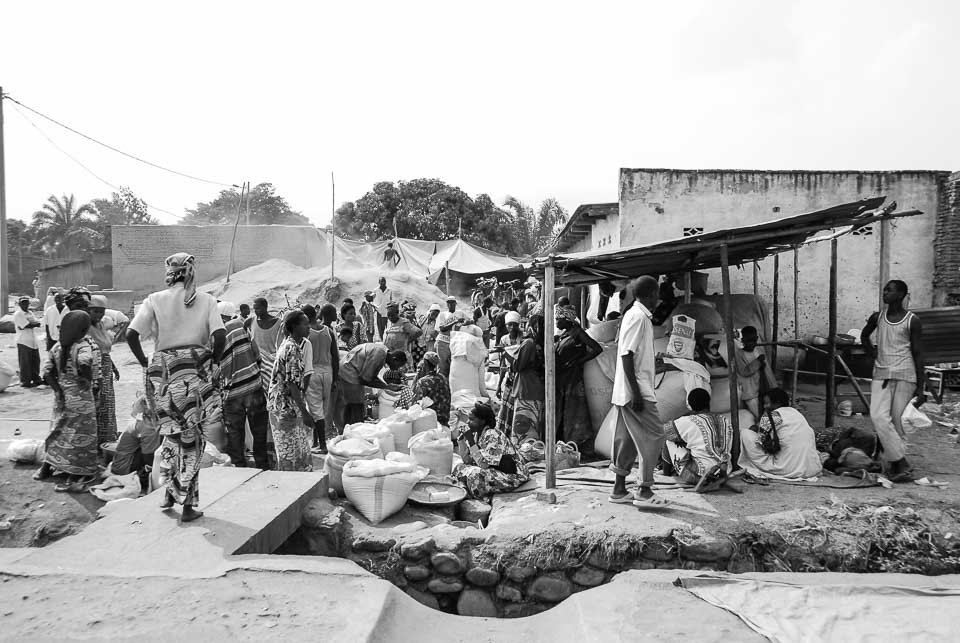 A busy outdoor marketplace with people gathered around bags and stalls in a rural area.
