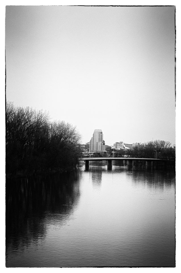 Black and white photo of a river with trees on the left, a bridge crossing over the river, and a city skyline with a tall building in the background.