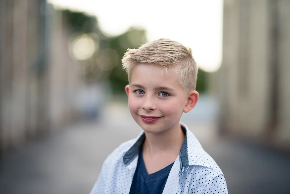 Close-up of a young boy with blonde hair, blue eyes, and a slight smile, standing outdoors on a blurred background.