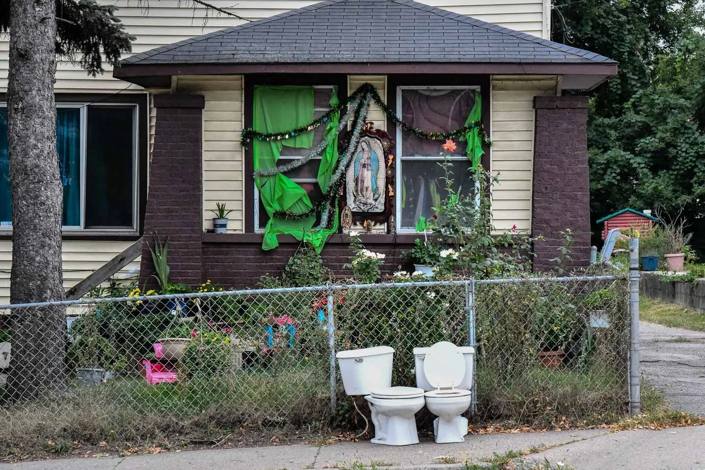 A house with a decorated front porch, black tinsel, and green fabric drapes. There is a picture of Our Lady of Guadalupe in the window, which is partly covered with curtains. Two toilets are placed outside in front of the yard, which is surrounded by