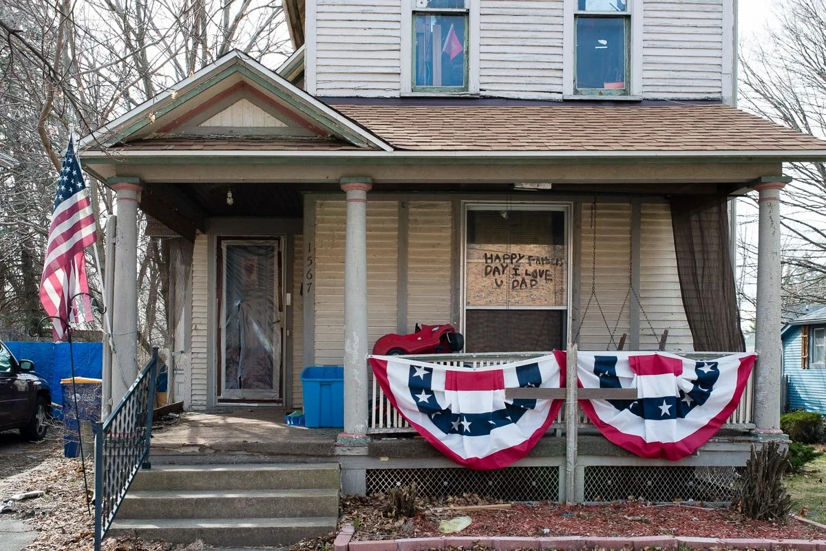 Front porch of a house decorated for a celebration, with American flag, patriotic bunting, a red toy car, and a handwritten sign in the window that says 'HAPPY FATHER'S DAY I LOVE U DAD.' The house has a screened-in porch, stairs leading up to the fr
