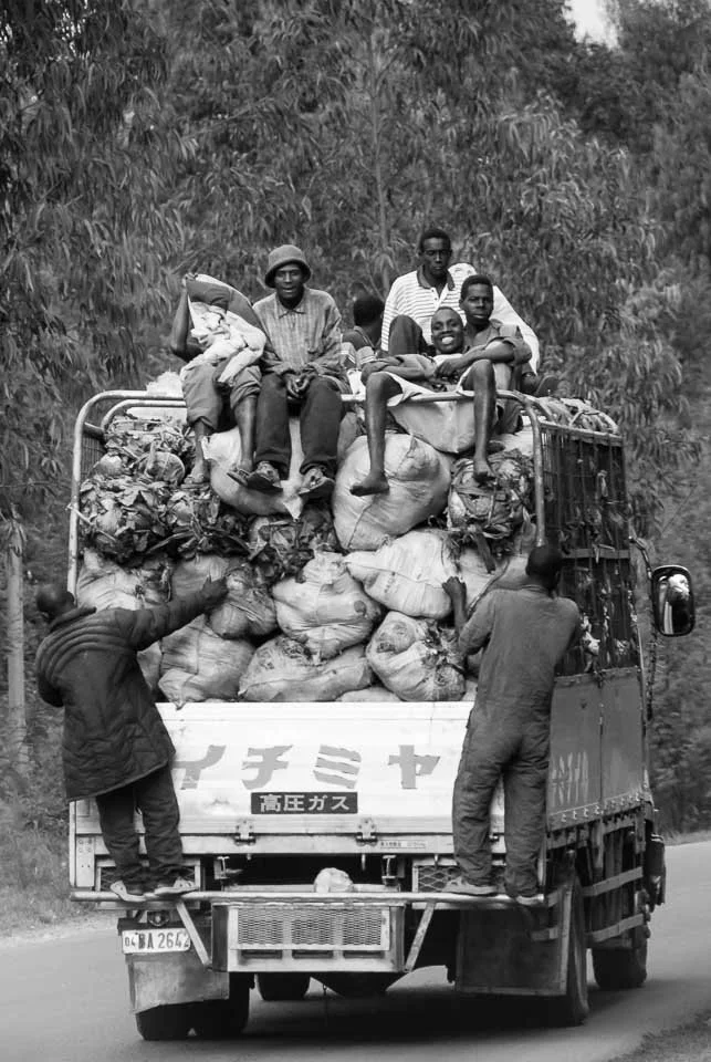 A truck loaded with sacks and bags, with several people sitting on top, being watched by two men hanging onto the back and sides. The truck is traveling on a rural road with trees in the background.