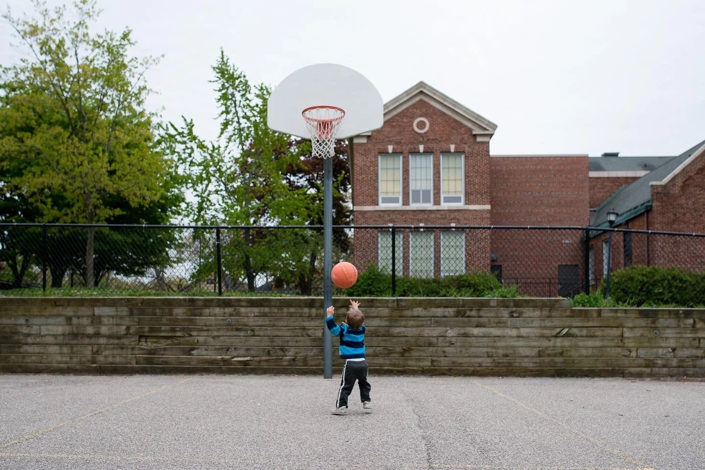 A young boy in a blue striped hoodie playing basketball on an outdoor court with a brick building in the background.