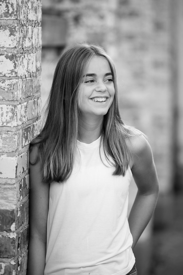 A young woman with long hair, smiling, leaning against a brick wall, in a sleeveless top, outdoors with a blurred background.