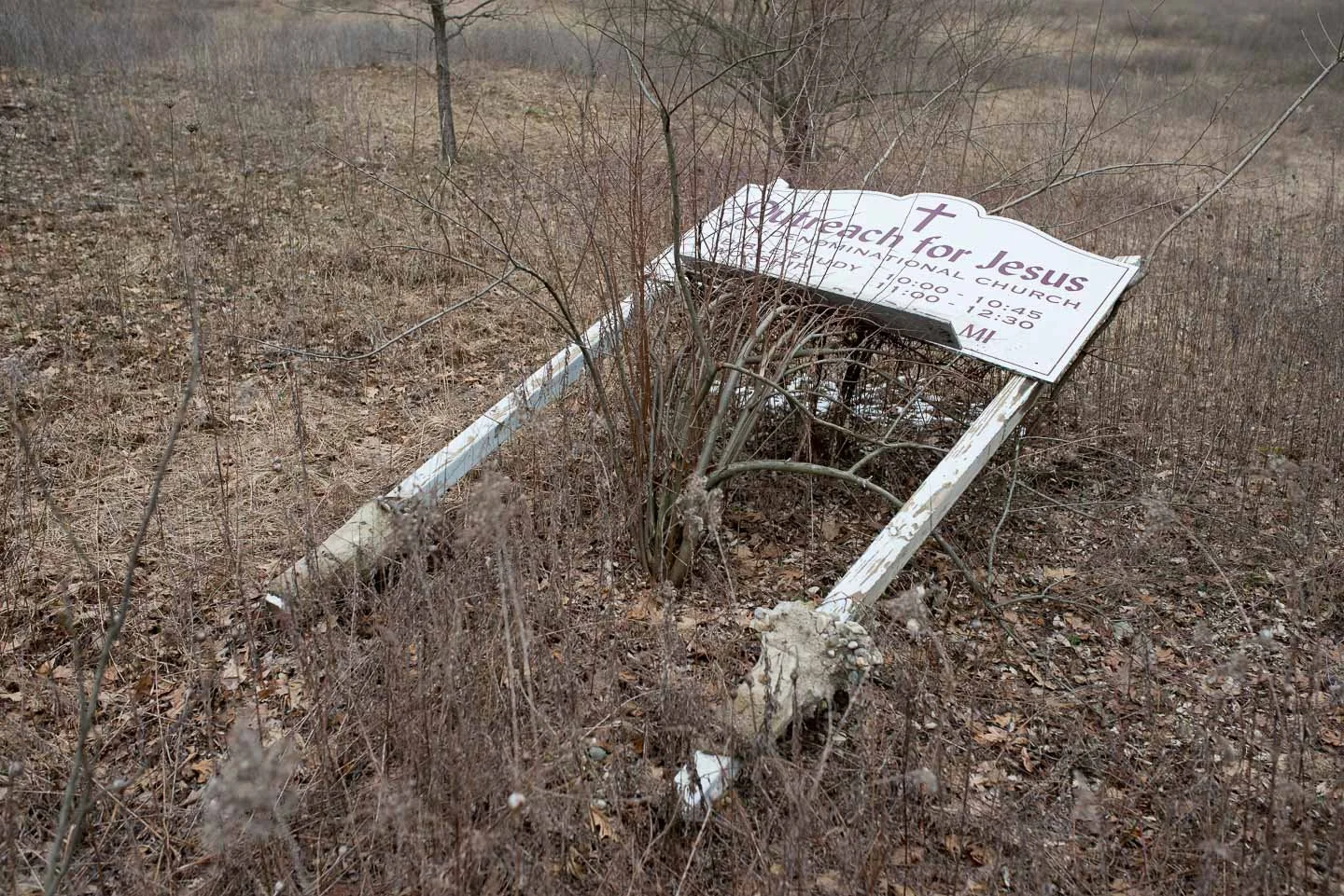 A fallen church sign for 'Reach for Jesus', lying on dry, brown land with weeds and small plants growing around it.