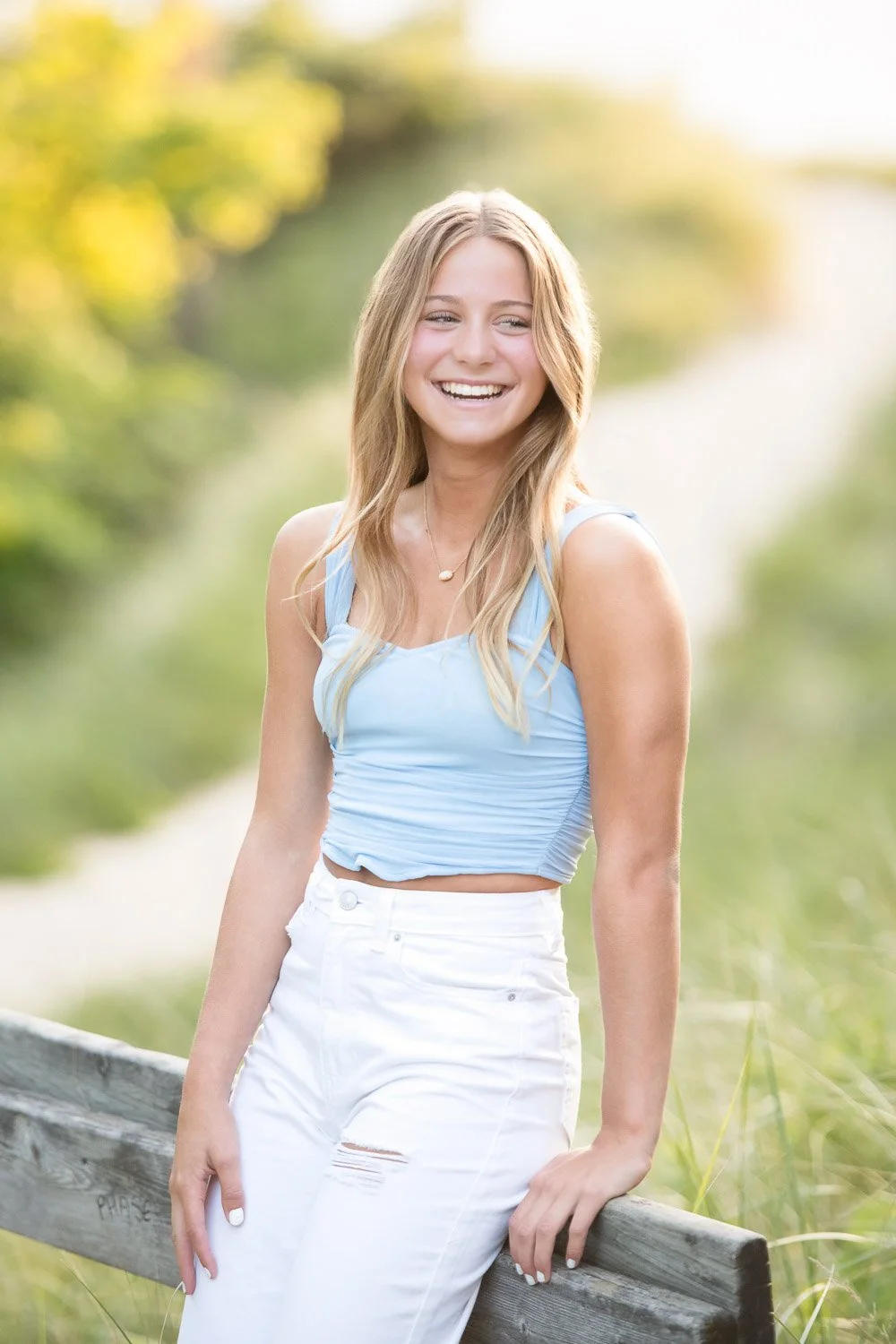 A young woman with long blonde hair smiling outdoors, wearing a light blue sleeveless crop top and white high-waisted distressed jeans, leaning against a wooden railing in a natural setting with blurred greenery in the background.