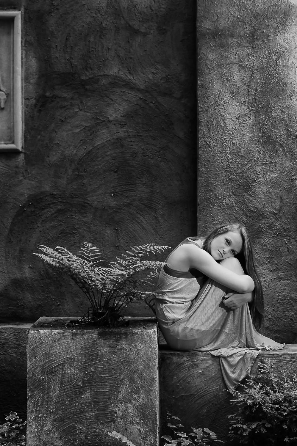 A black and white photograph of a young woman sitting on a stone ledge with her arms wrapped around her knees, looking thoughtfully into the camera. There are textured walls and plants around her.