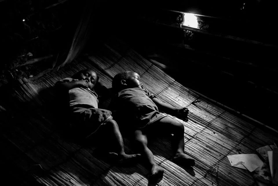 Two young children sleeping on a bamboo mat inside a dark room with a small opening letting in light.