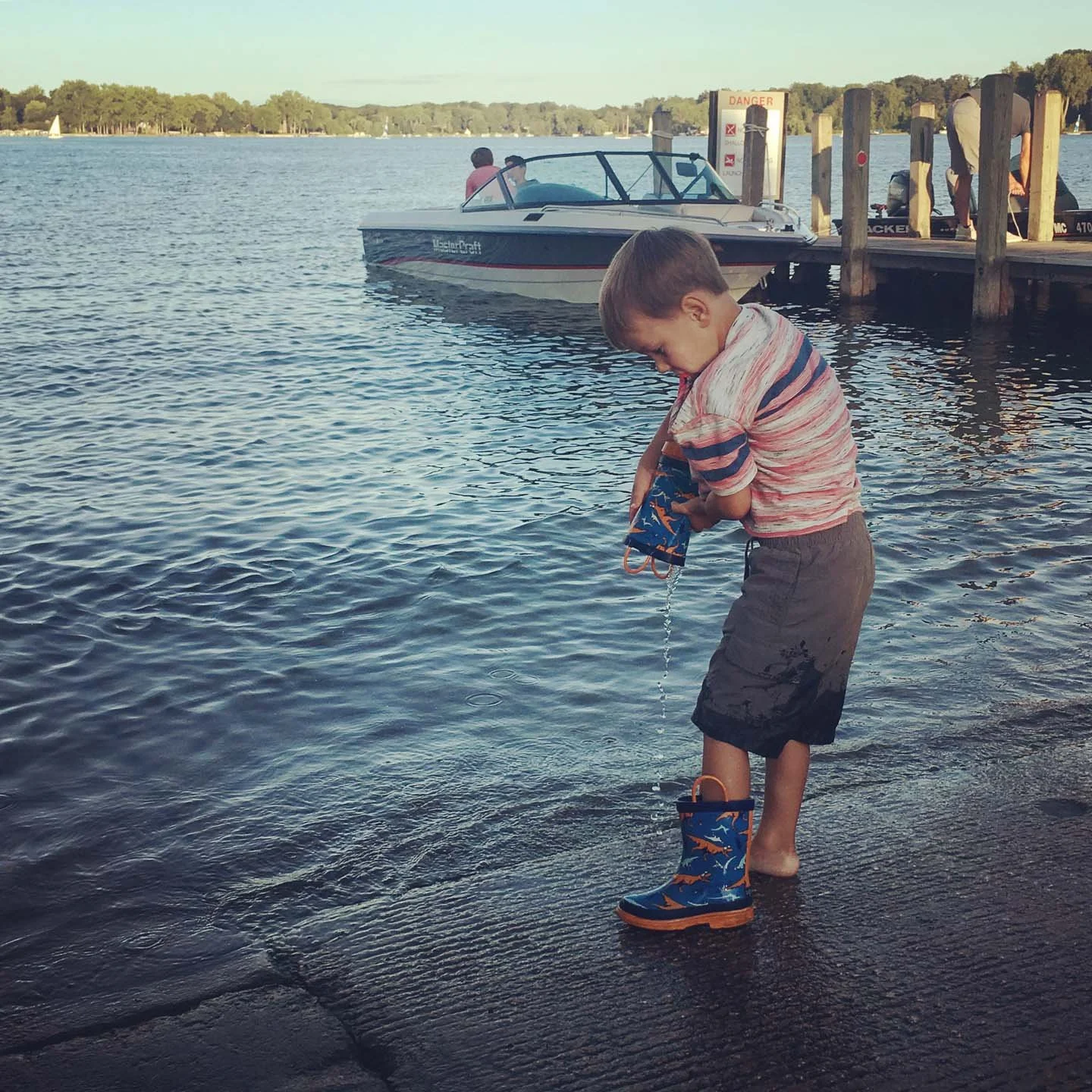 A young boy in striped shirt and shorts standing at the edge of a dock, holding a small blue bag with fish illustrations, while dipping his foot into the water near a boat on a lake.