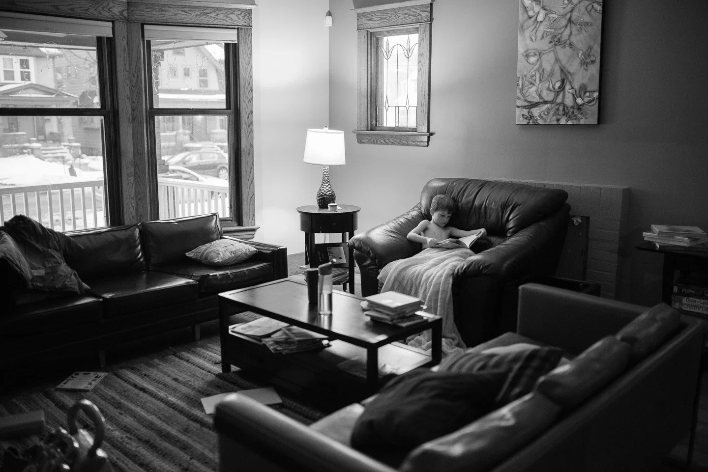 A black-and-white photo of a living room with a boy reading a book on a loveseat, next to a larger sofa with pillows. There are two windows showing a snowy exterior, a table with a lamp, and a coffee table with magazines. Artwork hangs on the wall, a