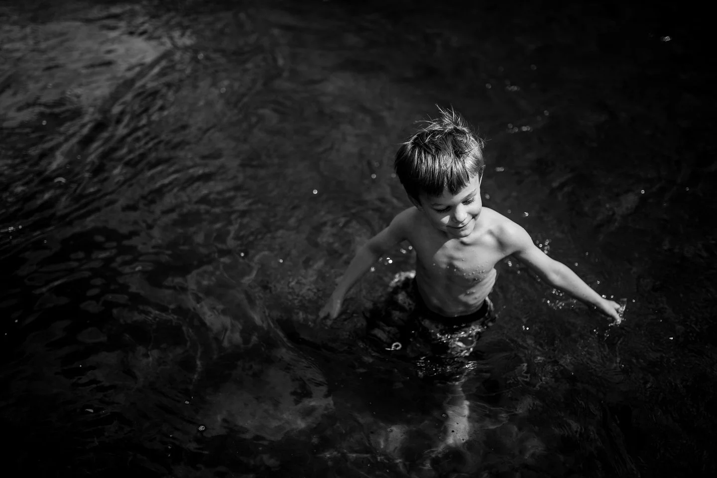 A young boy with short hair, shirtless, is enjoying himself in a body of water, smiling and splashing as he plays.