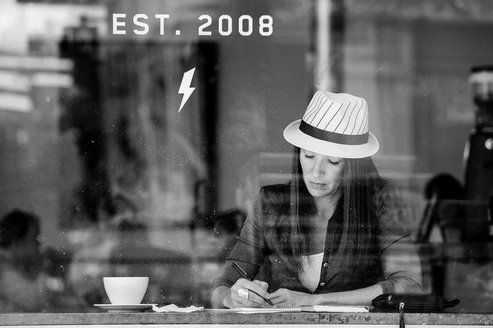 A woman sitting in a cafe, wearing a striped fedora hat, writing on a notepad, with a cup and some papers in front of her, seen through a glass window with reflection of the street outside.