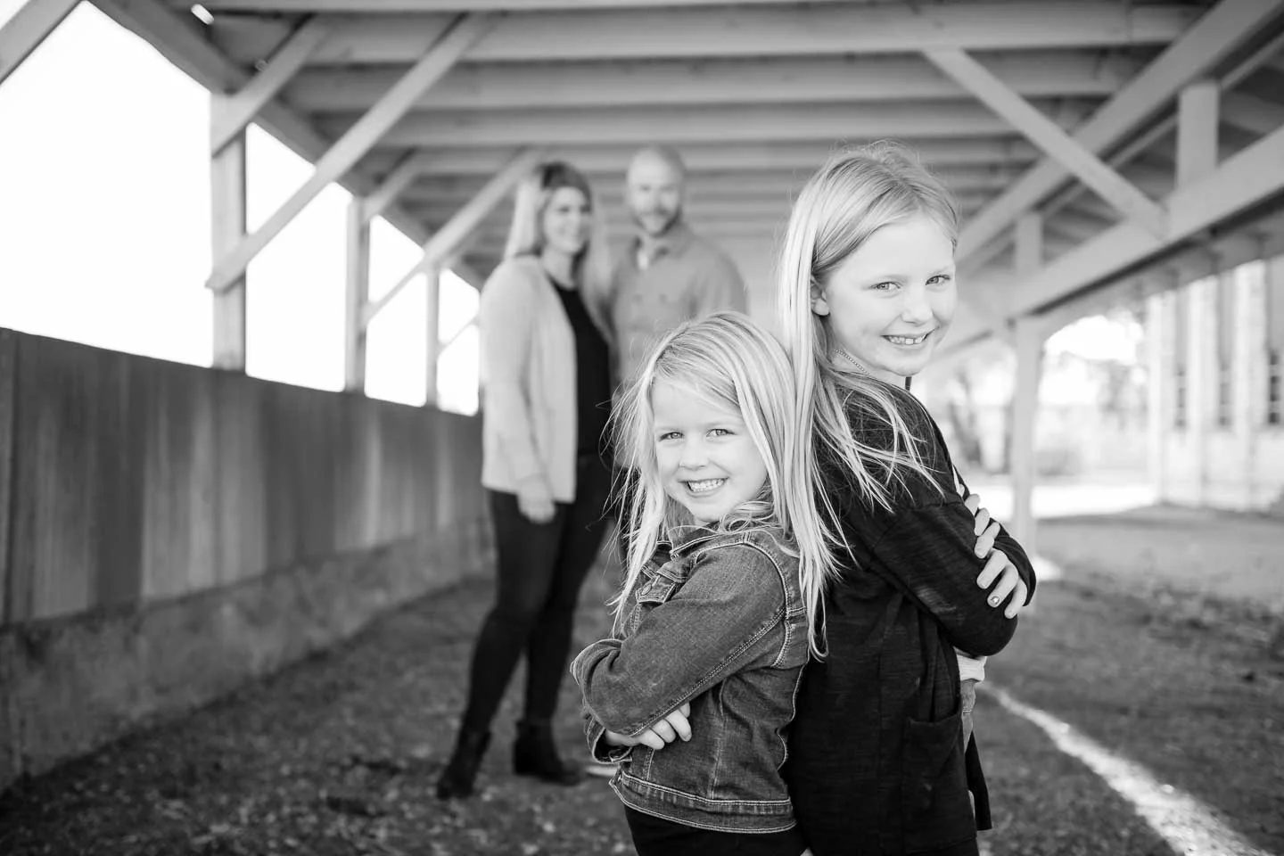 Two young girls stand in front of two adults in a covered outdoor structure, smiling and posing with crossed arms, with the adults blurred in the background.