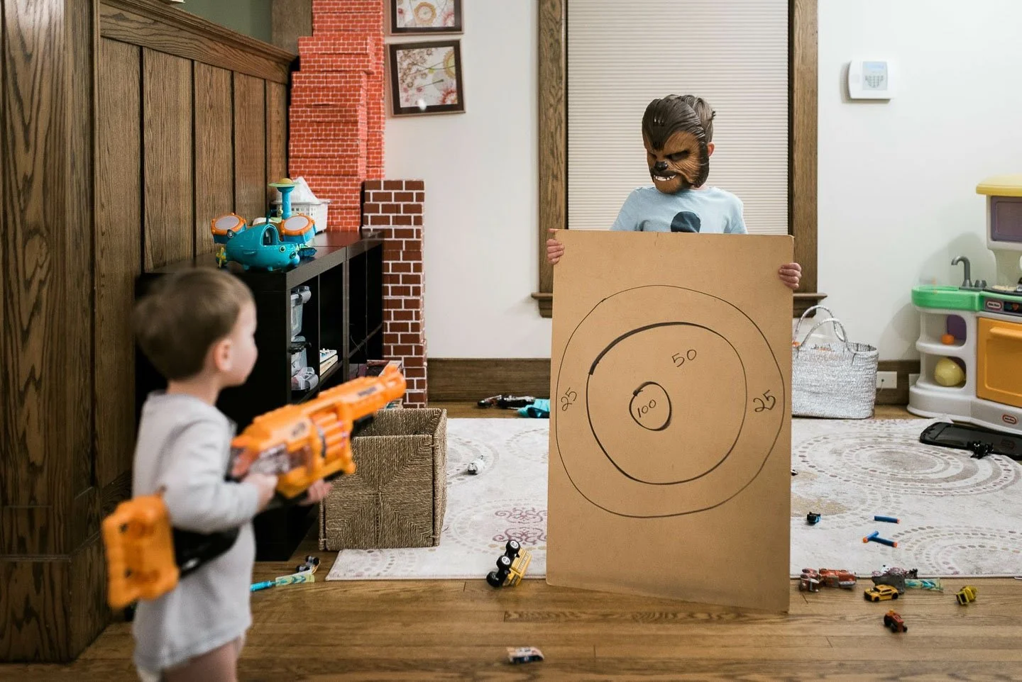 A child wearing a wolf mask is holding a cardboard target with concentric circles and numbers from 25 to 100, while another child in the foreground is aiming a toy gun. The scene is in a living room with toys scattered around.