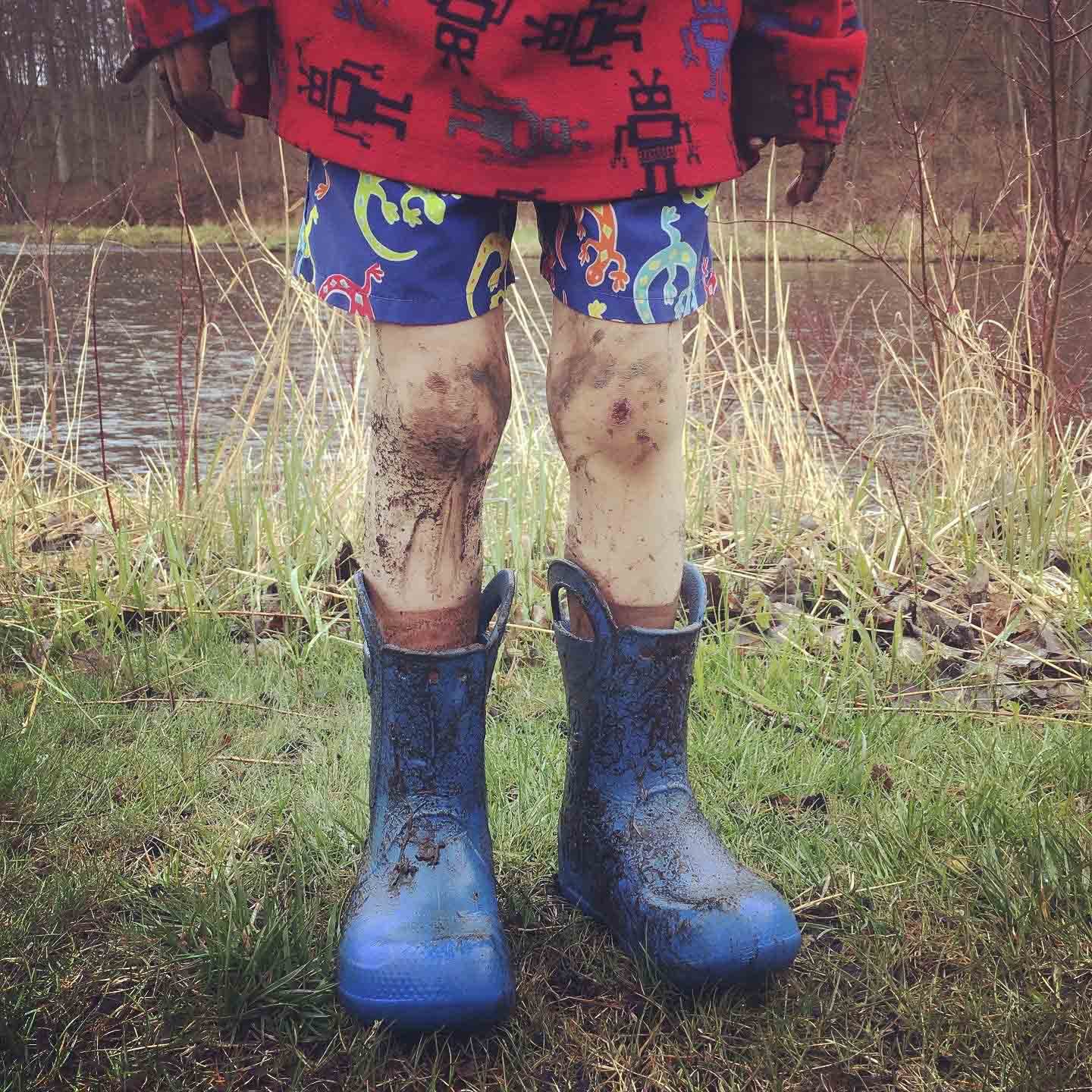 Child standing outdoors near a pond, wearing muddy blue rain boots, colorful shorts with dinosaur patterns, a red sweater with robot prints, and gloves, with muddy legs and grassy ground.
