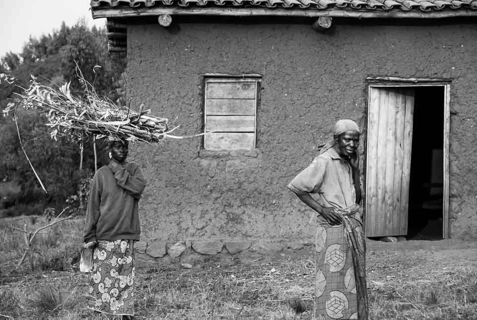An elderly man stands outside a simple mud house with a wooden door, smiling and holding a stick, while a woman carries a bundle of sticks on her head.