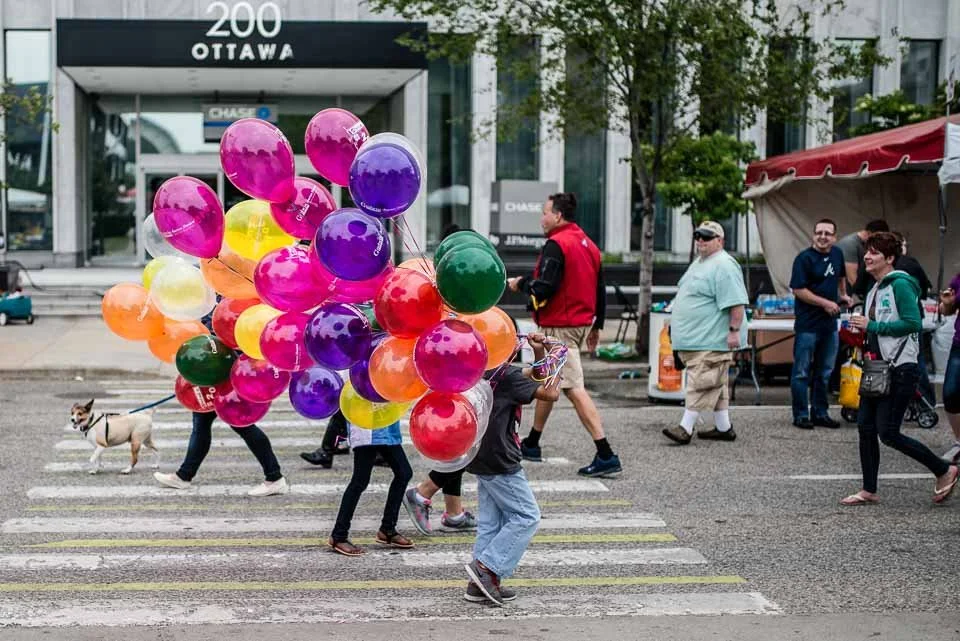 People crossing a city street at a crosswalk, with a person carrying numerous vibrant balloons in pink, purple, green, yellow, and orange colors.